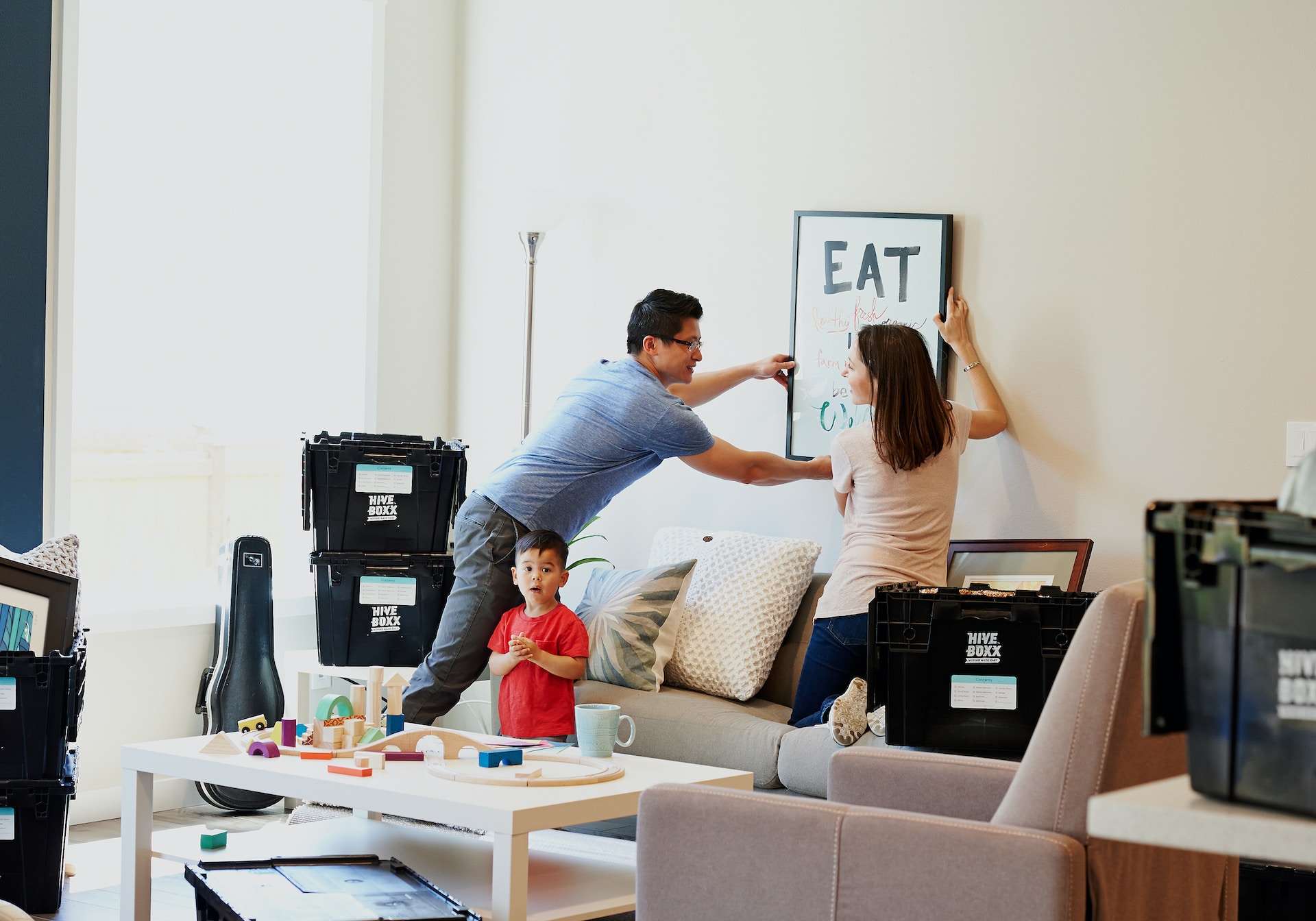 Mother and father putting a picture up on a wall surrounded by moving boxes. Toddler in front of them playing trains on a coffee table.