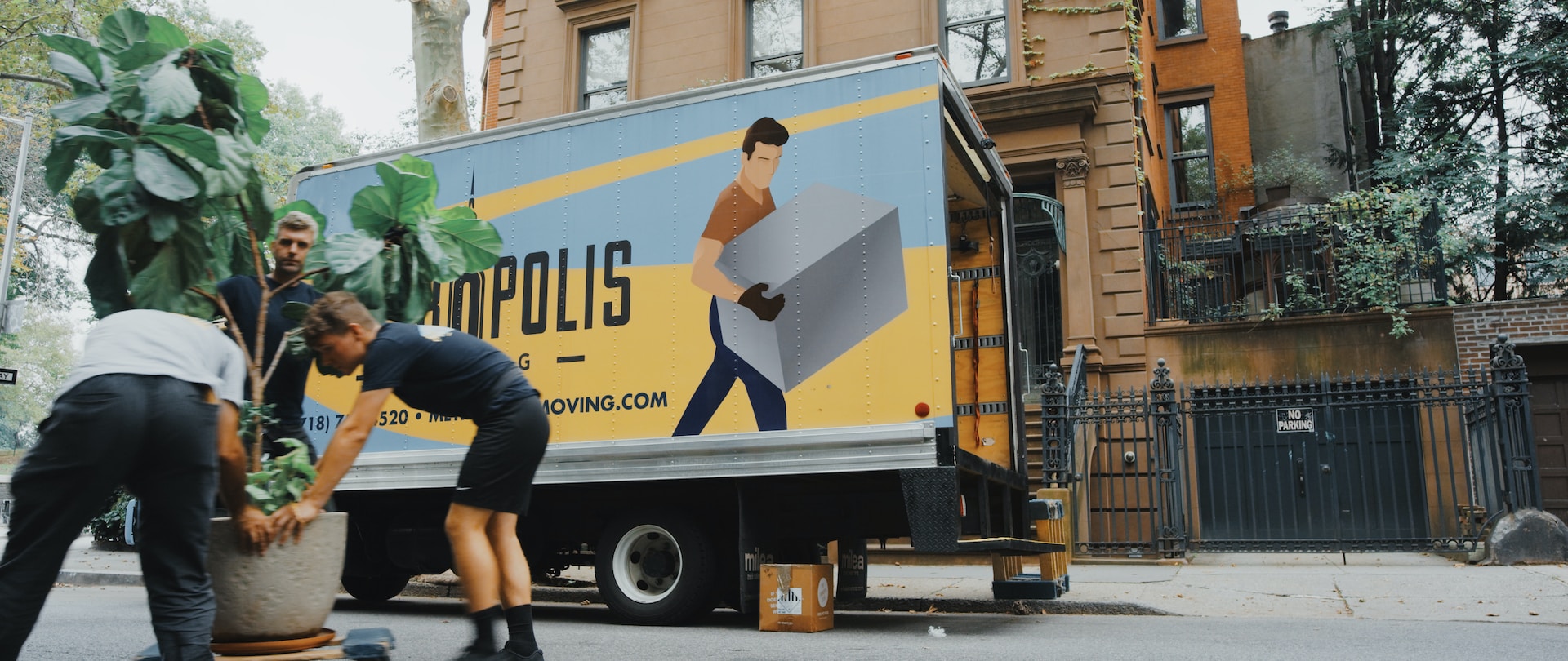 Moving van parked outside a residential building. Three men in foreground moving a large potted plant.